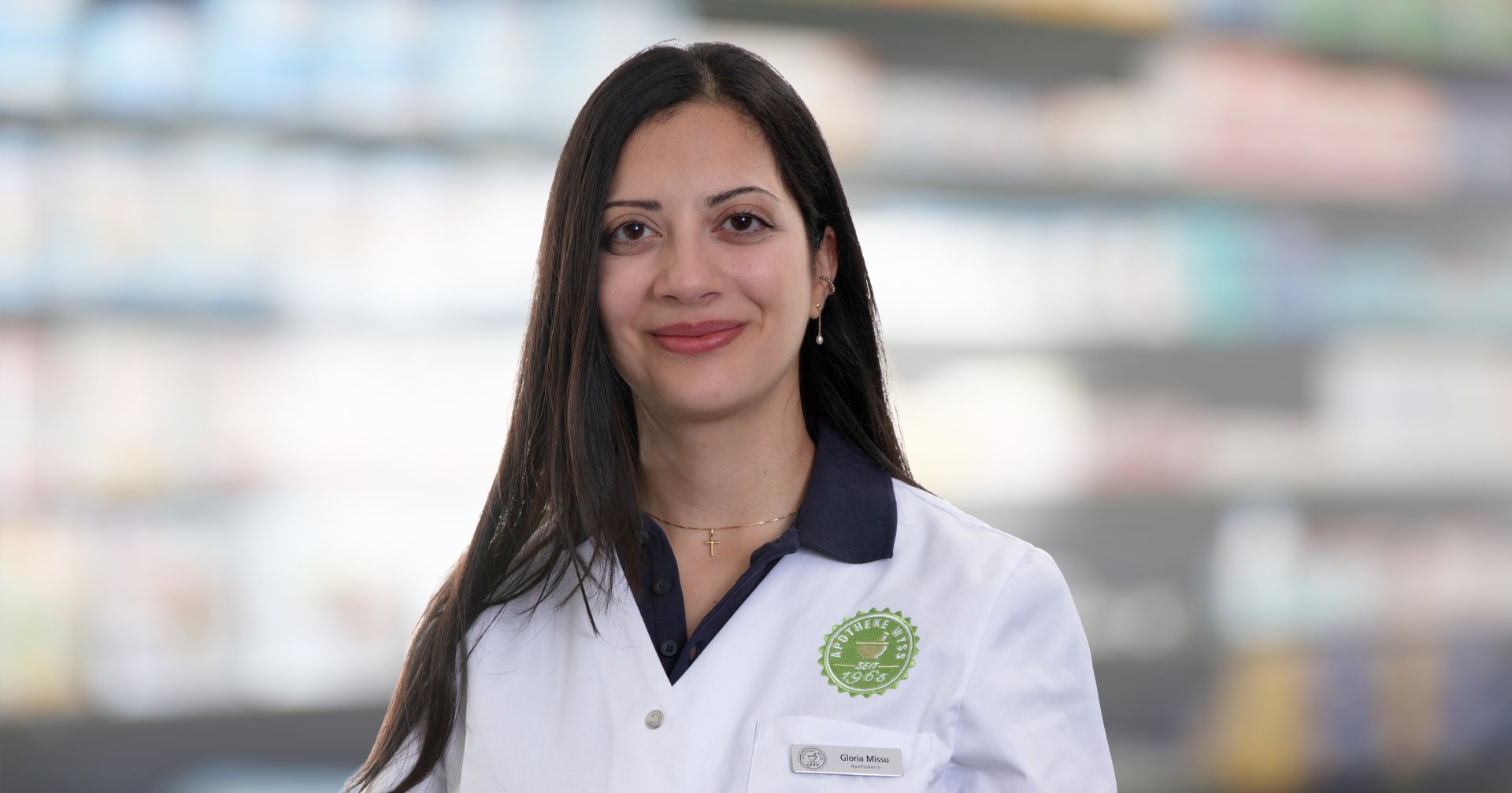 Joana Amaral Pedro, eine Frau mit langen dunklen Haaren, die einen weißen Apothekerkittel trägt, steht lächelnd vor verschwommenen Apothekenregalen.