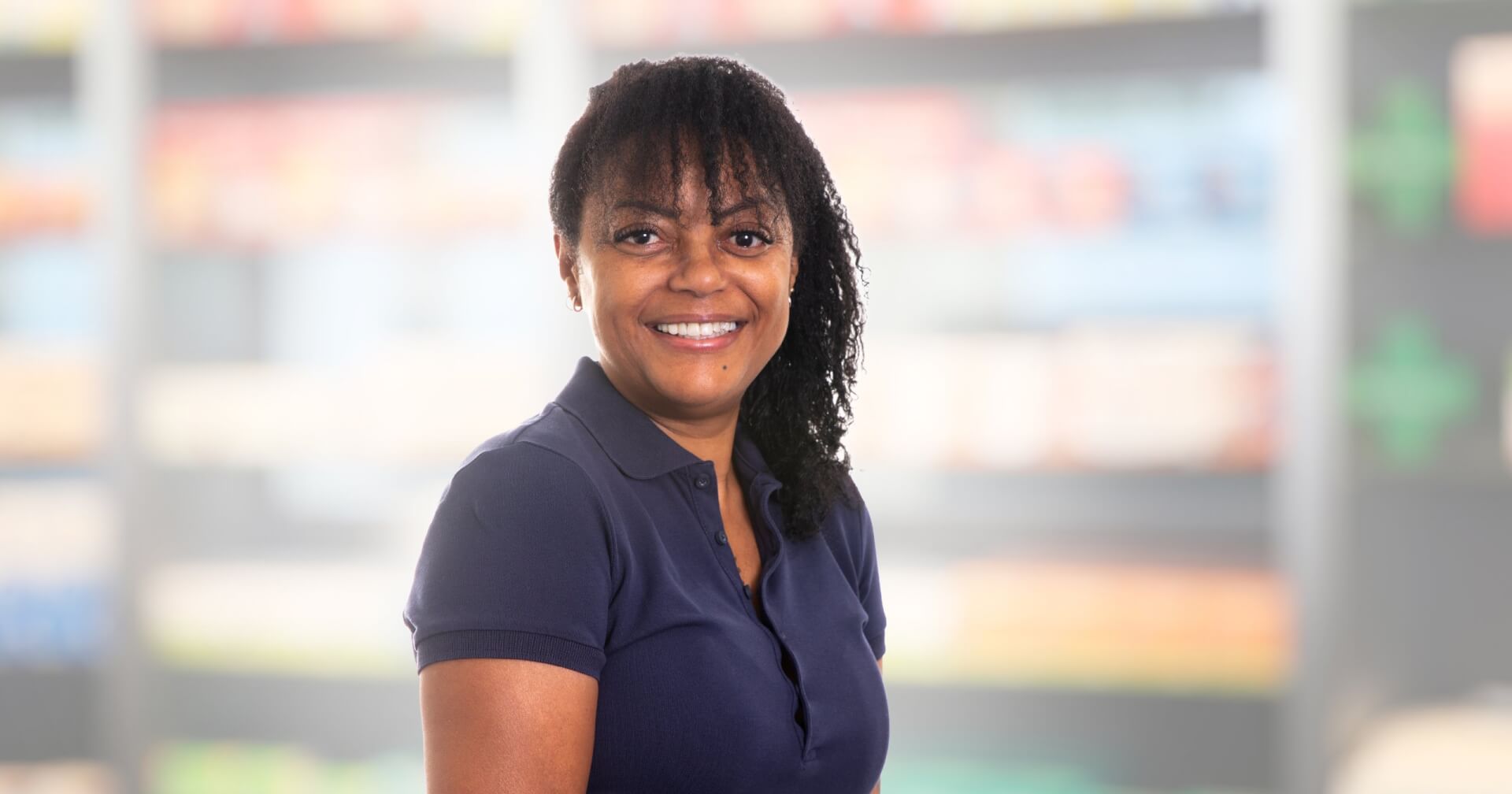Joana Amaral Pedro, eine Frau mit lockigem Haar und einem marineblauen Poloshirt, steht lächelnd vor einem unscharfen Hintergrund, der Regale in einem Geschäft oder einer Apotheke zu sein scheint.