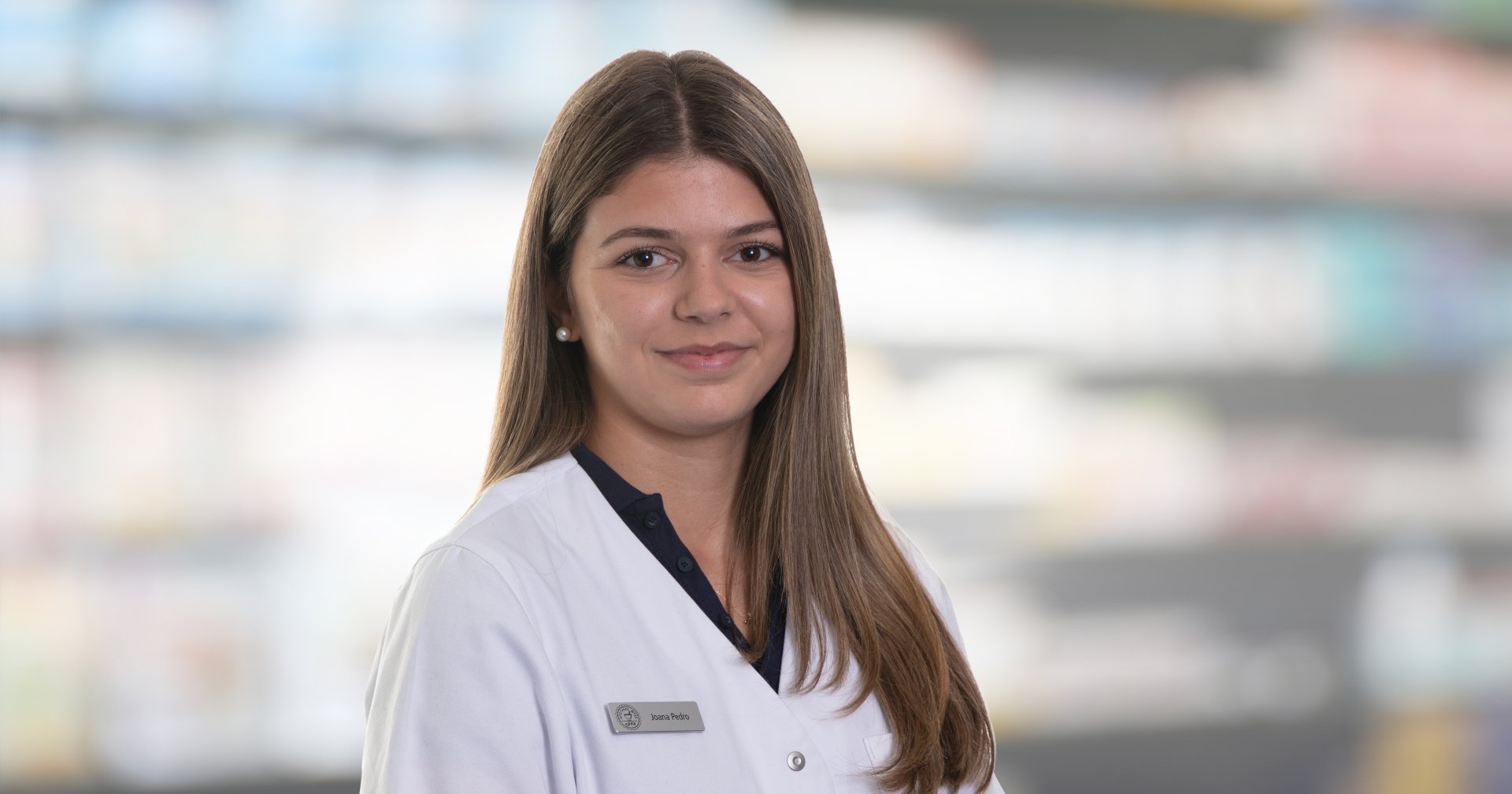 Joana Amaral Pedro, eine junge Frau mit langen braunen Haaren, trägt einen weißen Laborkittel und ein Namensschild, als sie lächelnd vor unscharfen Regalen in einer scheinbaren Apotheke oder medizinischen Einrichtung steht.