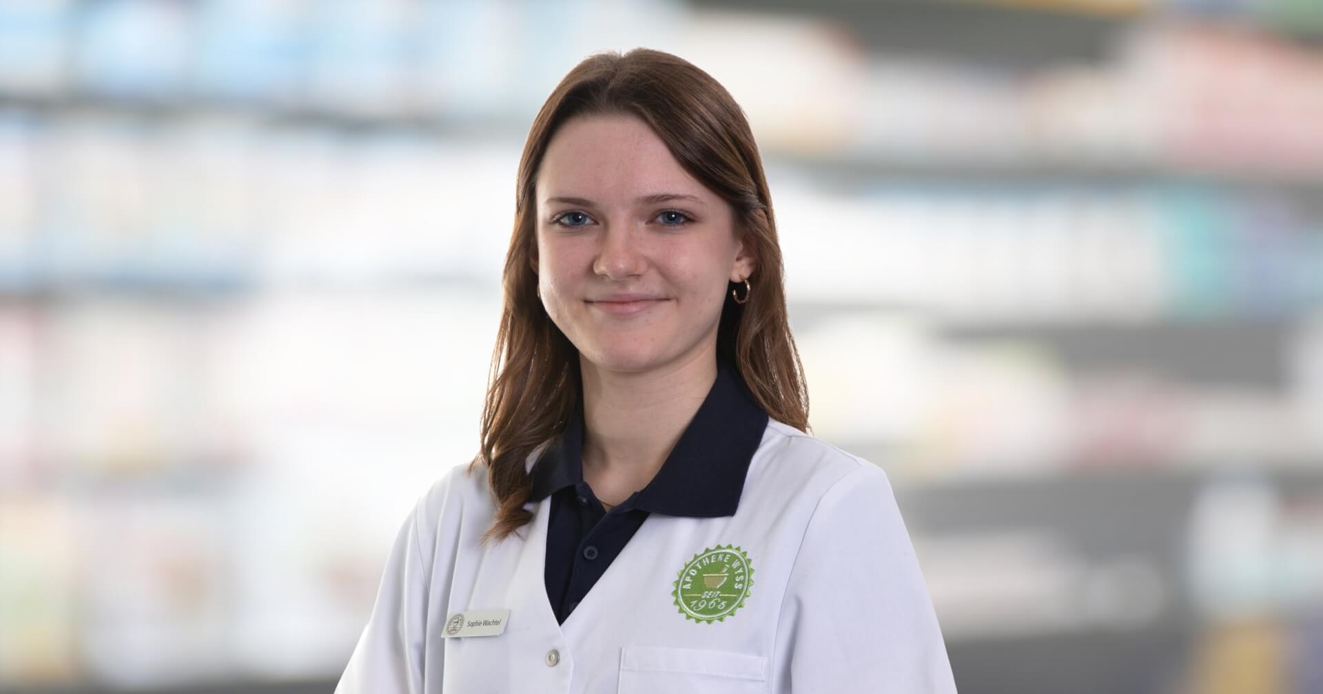 Joana Amaral Pedro, eine junge Frau mit weißem Laborkittel und Namensschild, steht lächelnd vor den verschwommenen Apothekenregalen.