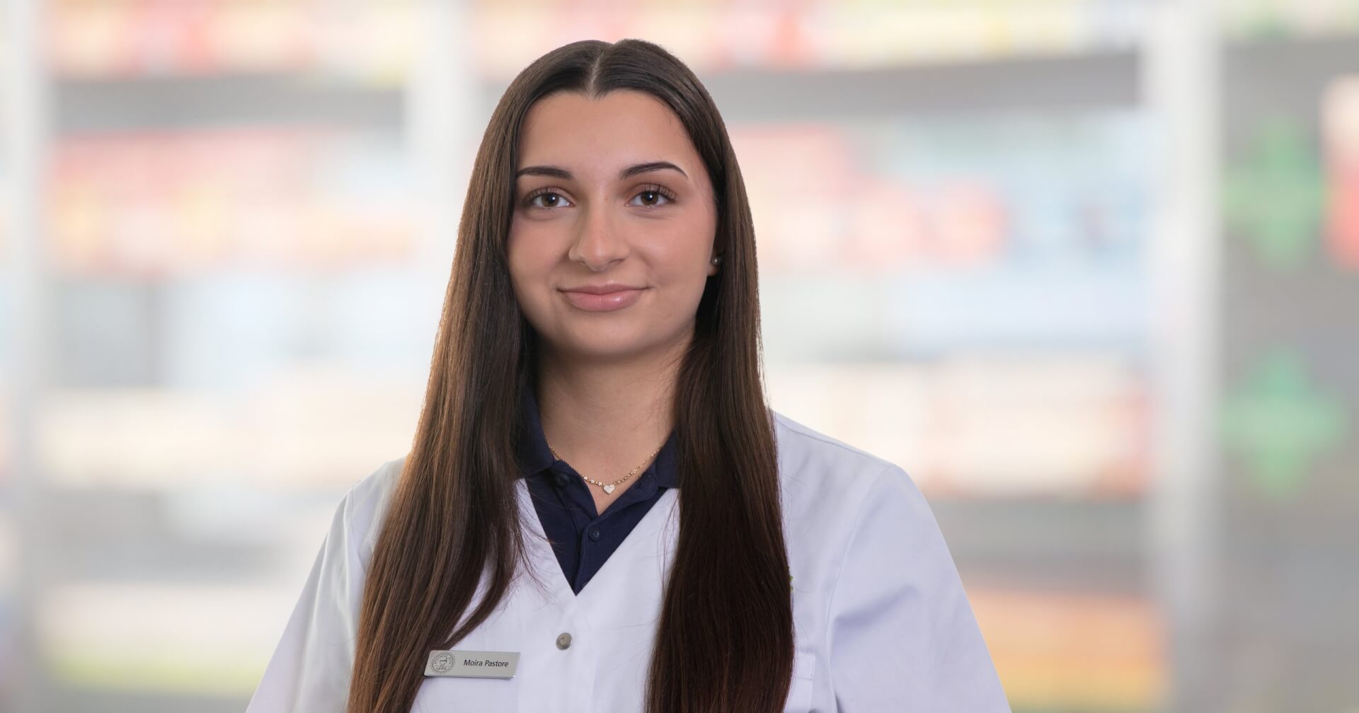 Joana Amaral Pedro, mit langen glatten braunen Haaren, weißem Laborkittel und Namensschild, steht vor unscharfen Apothekenregalen. Sie schaut in die Kamera und lächelt leicht.