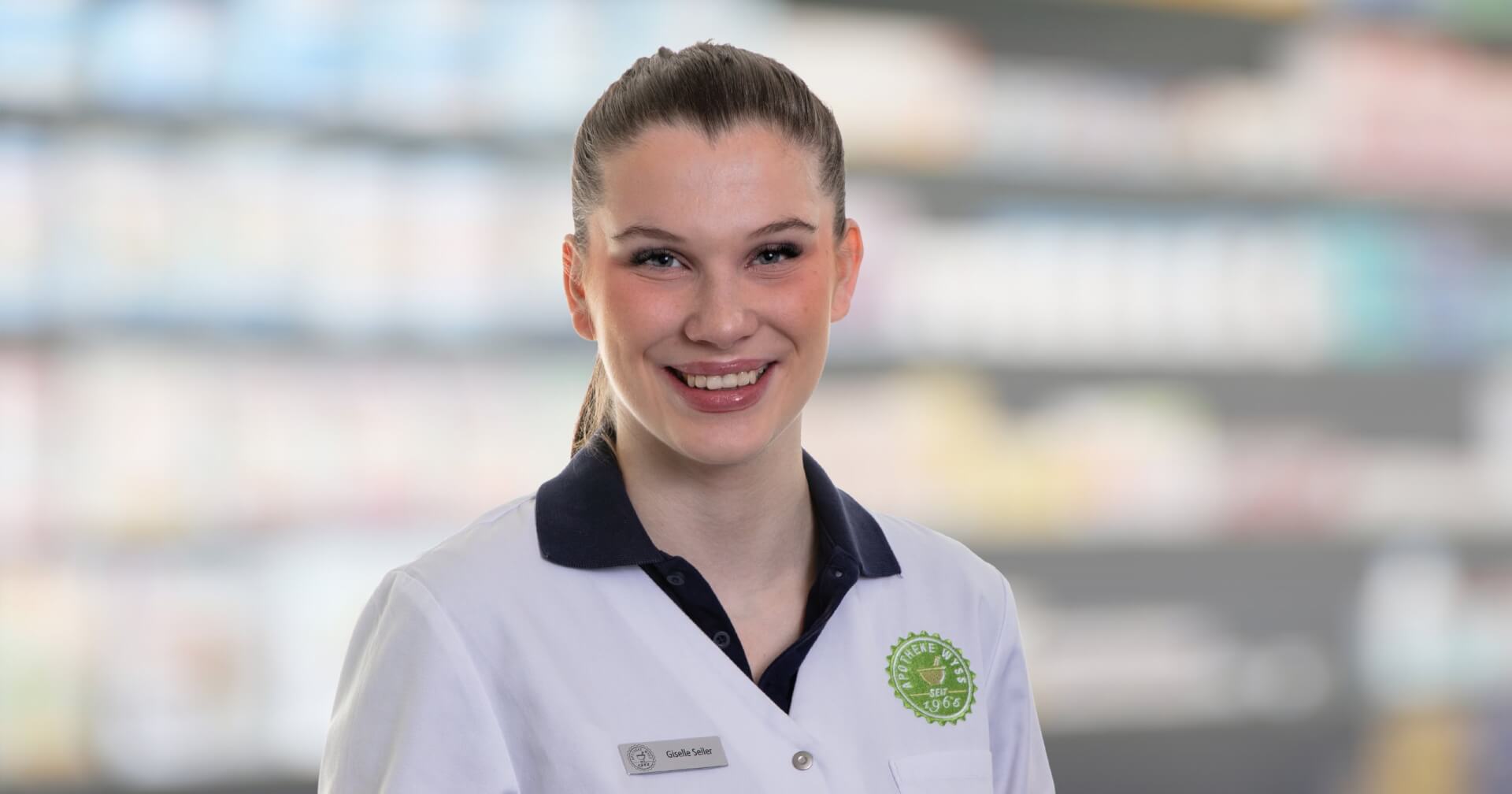 Eine lächelnde Frau, die durch ihr Namensschild als Joana Amaral Pedro identifiziert wird, trägt einen weißen Laborkittel, während sie vor verschwommenen Apothekenregalen steht, was darauf hindeutet, dass sie Apothekerin oder medizinisches Fachpersonal ist.