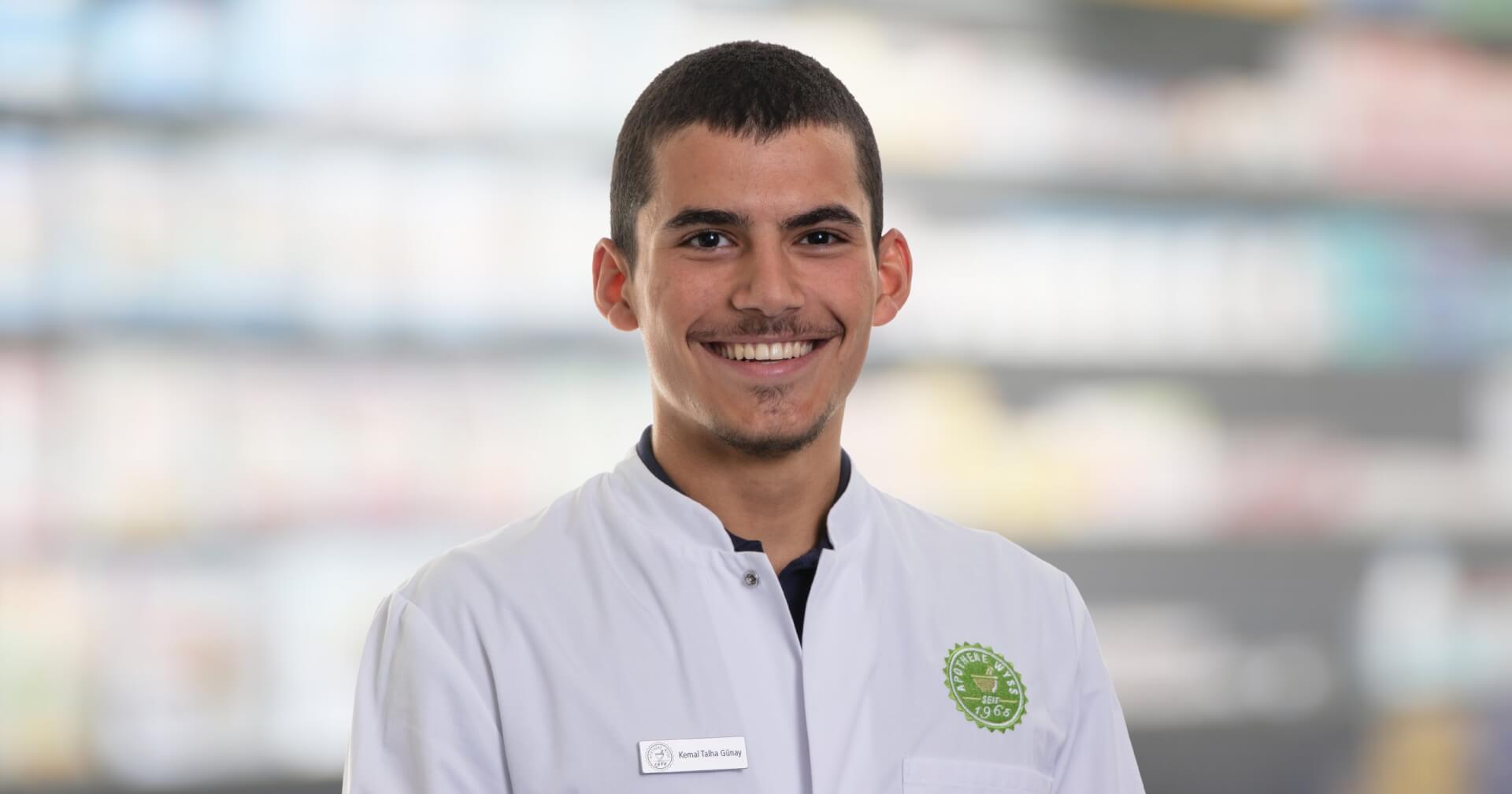 Ein junger Mann, der einen weißen Laborkittel mit einem grünen Emblem und einem Namensschild mit der Aufschrift Joana Amaral Pedro trägt, lächelt in die Kamera. Der unscharfe Hintergrund deutet auf einen Innenraum hin, möglicherweise eine Apotheke oder ein Labor.