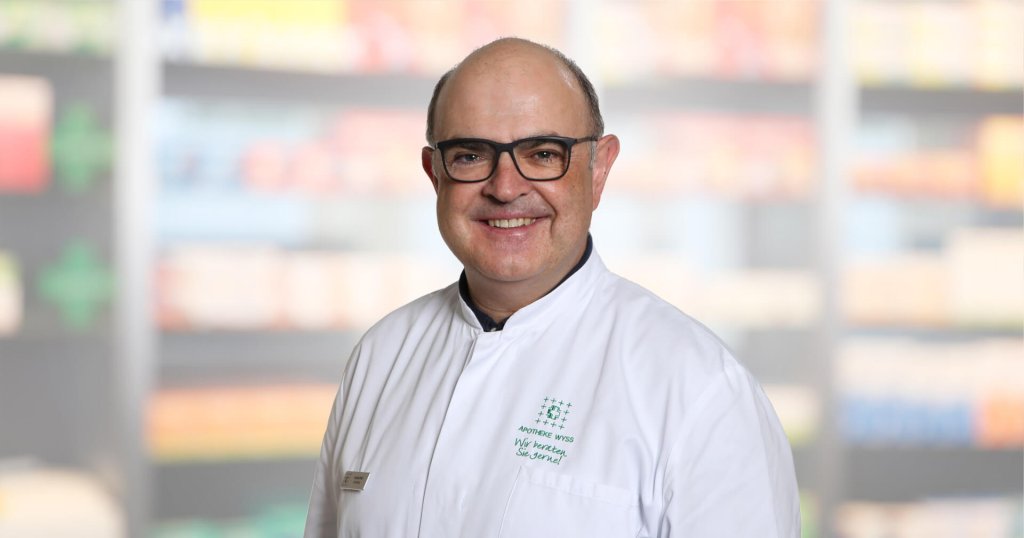 Ein lächelnder Mann mit Brille und weißem Laborkittel steht vor verschwommenen Regalen, wahrscheinlich in einer Apotheke oder in einem medizinischen Bereich.