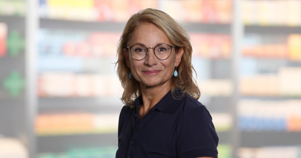 Helen Hagmann, eine Frau mittleren Alters mit blondem Haar, Brille und blauen Ohrringen, lächelt und trägt ein marineblaues Poloshirt. Sie steht vor verschwommenen Regalen, möglicherweise in einem Geschäft oder einer Apotheke.