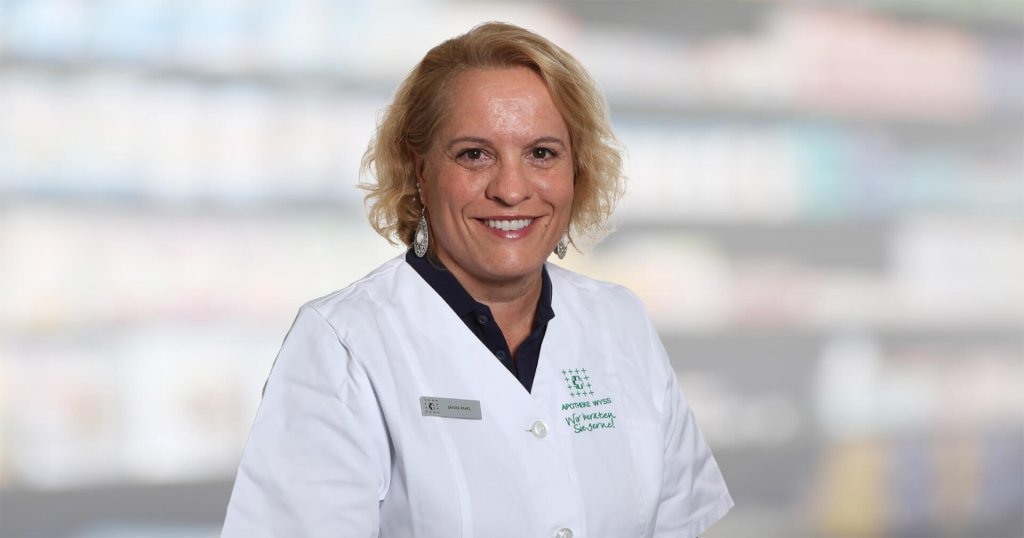 Milica Dašić, eine Frau mit blondem Haar, lächelt, während sie einen weißen Laborkittel und ein Namensschild trägt. Sie steht vor unscharfen Regalen, wahrscheinlich in einer Apotheke oder einem medizinischen Bereich.