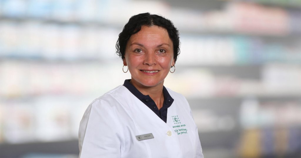 Silvia Röder, eine Frau mit kurzen dunklen Haaren, die einen weißen Laborkittel und ein Namensschild trägt, lächelt in die Kamera. Der unscharfe Hintergrund deutet auf Regale oder eine Apothekeneinrichtung hin.