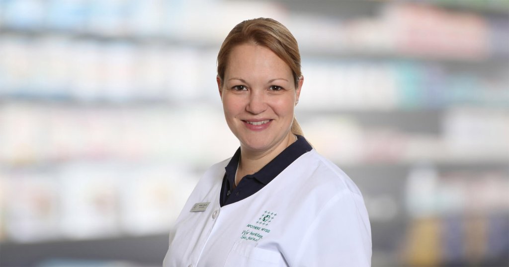 Mariana Schaller, eine lächelnde Frau in einem weißen Laborkittel, steht vor verschwommenen Regalen, die an eine Apotheke oder einen medizinischen Bereich erinnern.