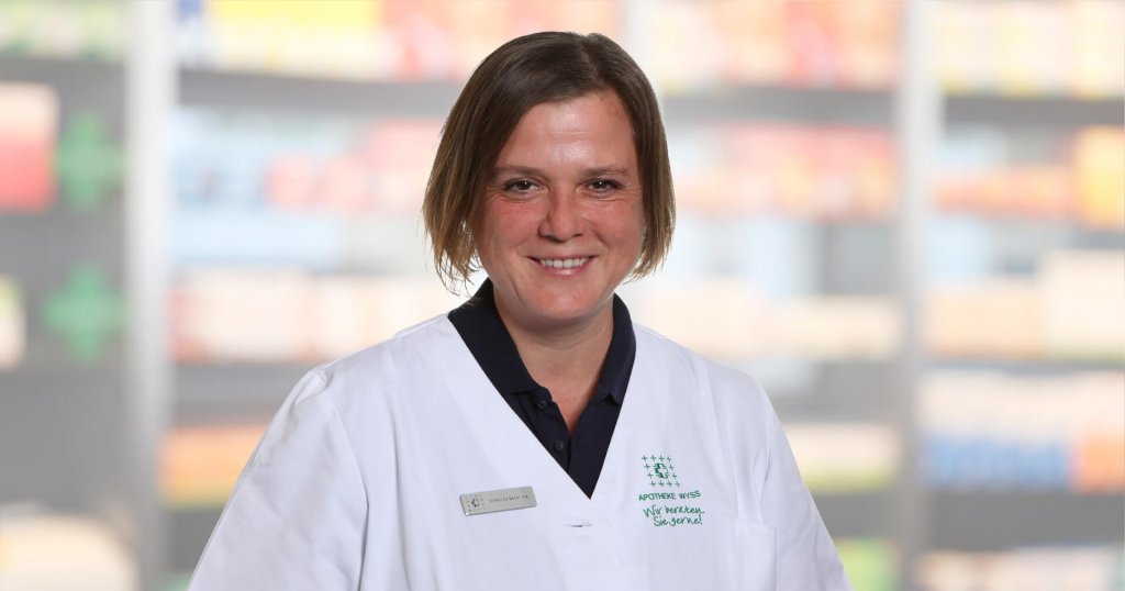 Natascha Reinhard, eine Frau mit kurzen braunen Haaren, die einen weißen Laborkittel und ein Namensschild trägt, lächelt, während sie vor unscharfen Regalen steht, möglicherweise in einer Apotheke oder einer medizinischen Einrichtung.