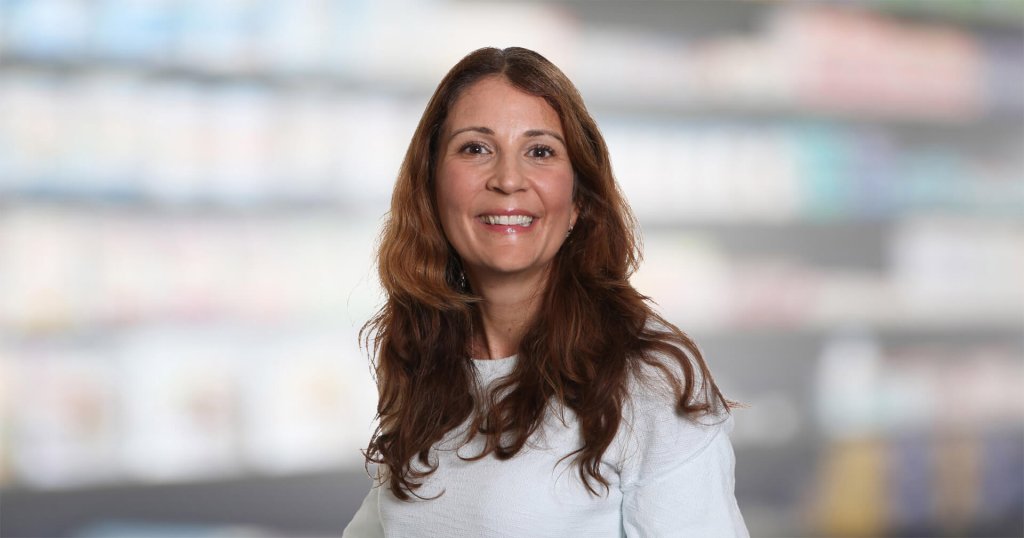 Magdalena Doukas, eine Frau mit langen braunen Haaren und einem weißen Oberteil, lächelt in die Kamera, während sie vor einem unscharfen Innenhintergrund mit Regalen steht.