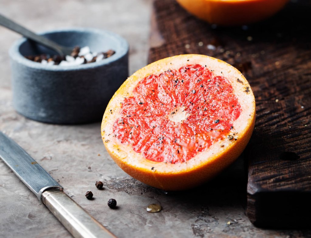 Eine halbierte, bittere und doch gesunde Grapefruit, bestreut mit Pfeffer, liegt auf einer rustikalen Fläche neben einem Holzbrett, einem Messer, Pfefferkörnern und einer kleinen Schale mit Salz und einem Löffel.