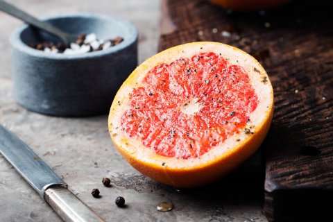 Eine halbierte, bittere und doch gesunde Grapefruit, bestreut mit Pfeffer, liegt auf einer rustikalen Fl&auml;che neben einem Holzbrett, einem Messer, Pfefferk&ouml;rnern und einer kleinen Schale mit Salz und einem L&ouml;ffel.