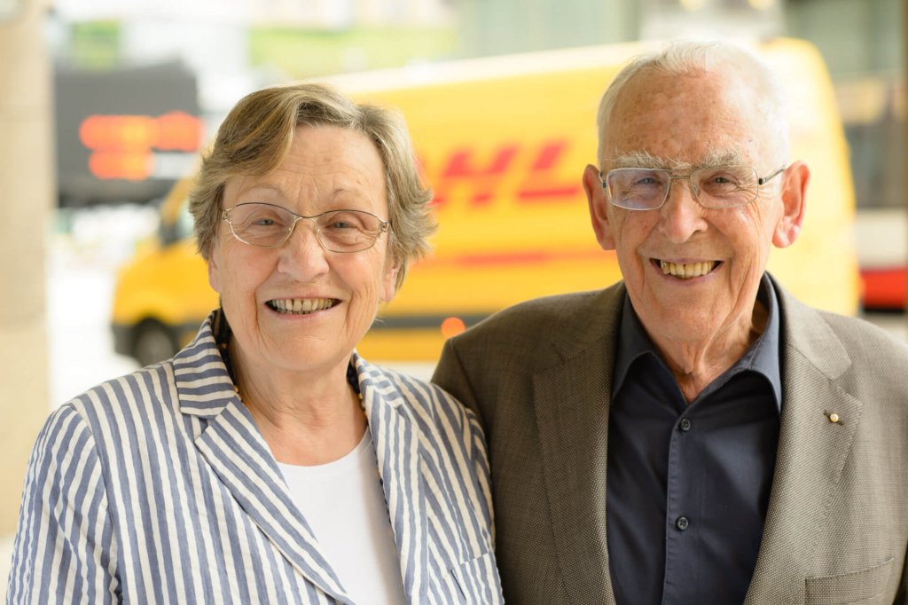 Eine ältere Frau und ein älterer Mann lächeln im Freien in die Kamera, im Hintergrund ist ein gelber DHL-Lieferwagen zu sehen. Die Frau trägt eine Brille und eine gestreifte Jacke; der Mann trägt eine Brille und einen braunen Blazer.