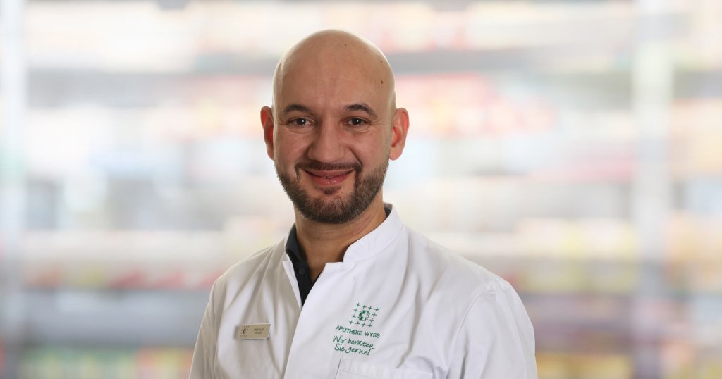 Ein lächelnder Mann mit Glatze und gestutztem Bart trägt einen weißen Arztkittel und steht vor einem unscharfen Hintergrund mit Regalen.