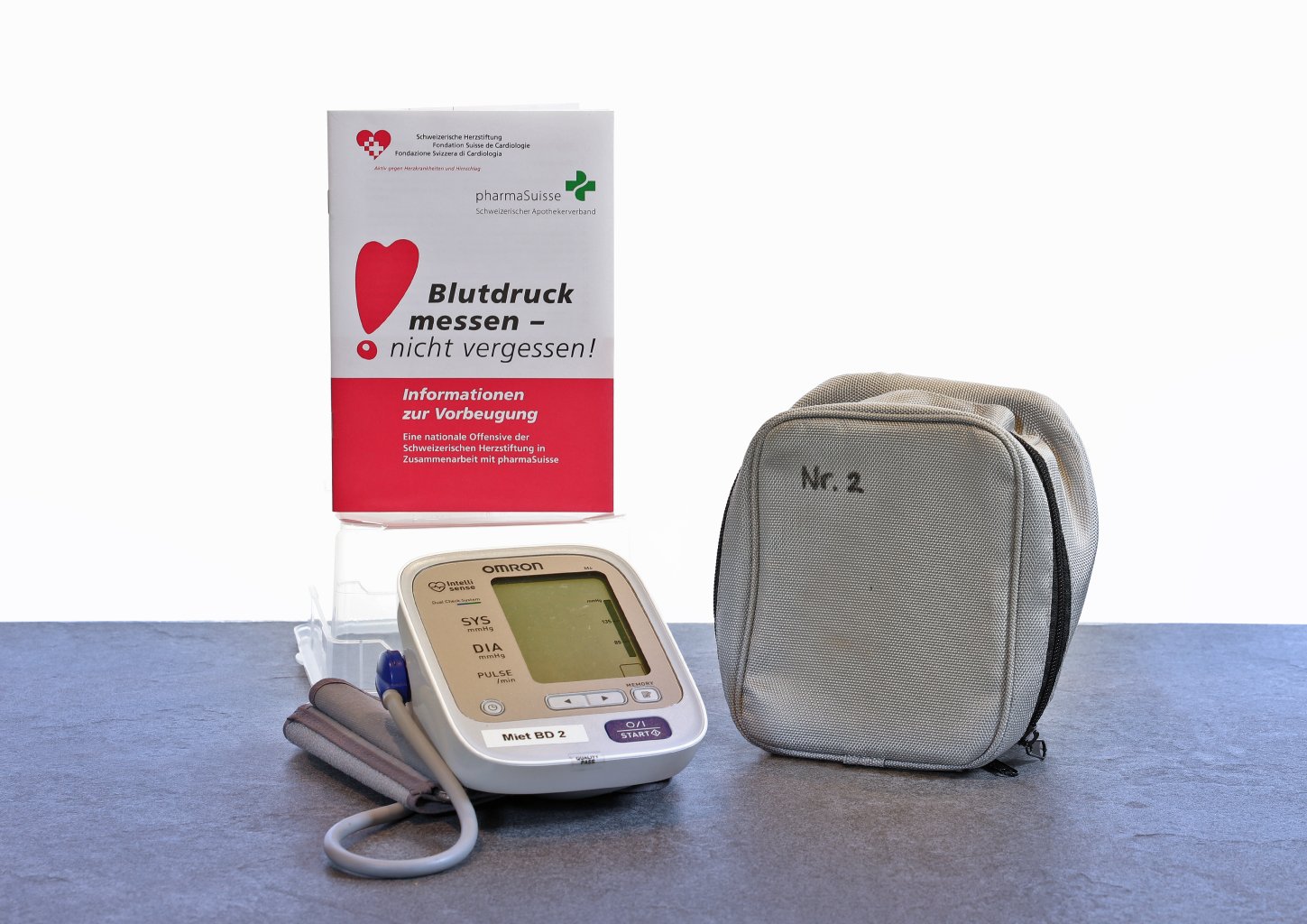 Ein Blutdruckmessger&auml;t mit Manschette und eine graue Tragetasche mit der Aufschrift Nr. 2 liegen auf einem Tisch. Dahinter liegt eine deutsche Brosch&uuml;re &uuml;ber Blutdruckmessung und Pr&auml;ventionsinformationen.