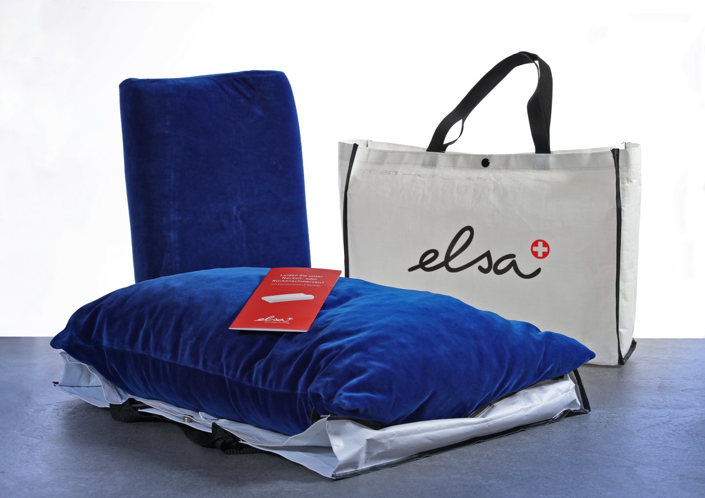 Ein blaues Kissen und eine blaue R&uuml;ckenlehne, die mit einem roten Etikett versehen sind, stehen als Sanit&auml;tsartikel vor einer wei&szlig;en elsa-Tasche mit schwarzen Griffen, alles auf einer grauen Fl&auml;che vor einem wei&szlig;en Hintergrund.