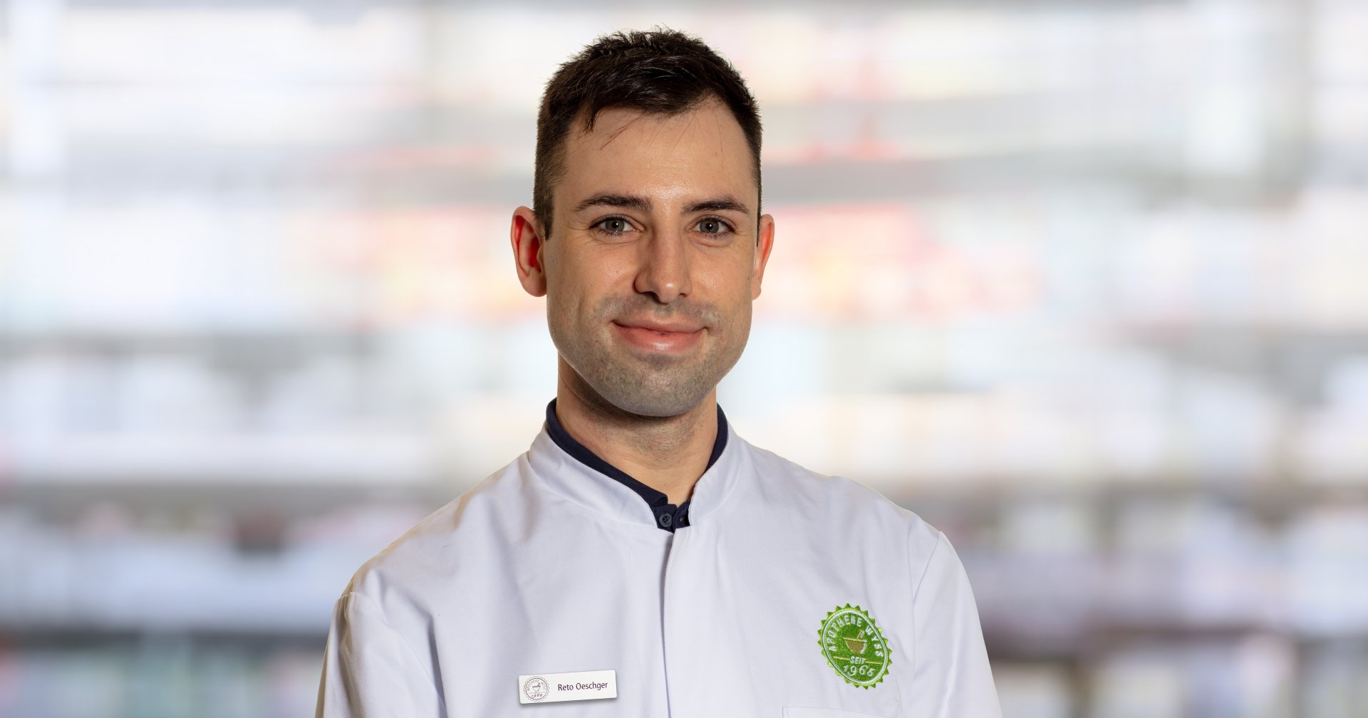 Ein Mann mit kurzen dunklen Haaren, der eine weiße Kochjacke mit einem grünen kreisförmigen Logo und einem Namensschild mit der Aufschrift "Reto Oeschger" trägt, steht vor einem verschwommenen Innenhintergrund und lächelt leicht in die Kamera.