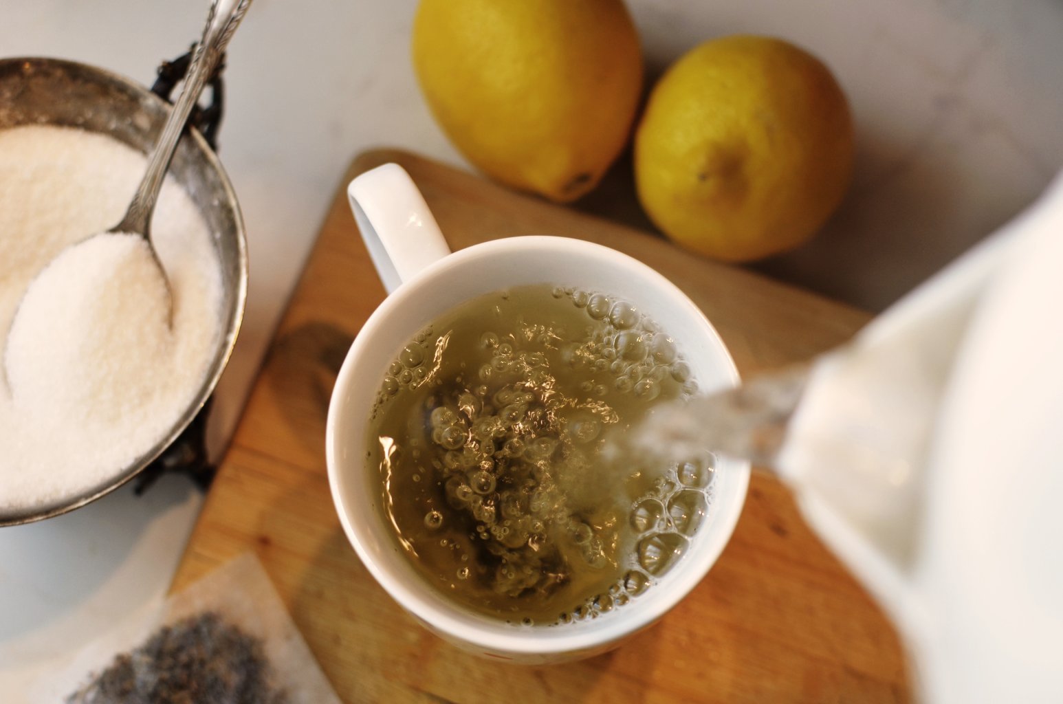 Eine wei&szlig;e Tasse wird auf einem Holzbrett mit hei&szlig;em Wasser f&uuml;r Tee gef&uuml;llt, daneben liegen zwei Zitronen, ein L&ouml;ffel Zucker und ein Teebeutel.