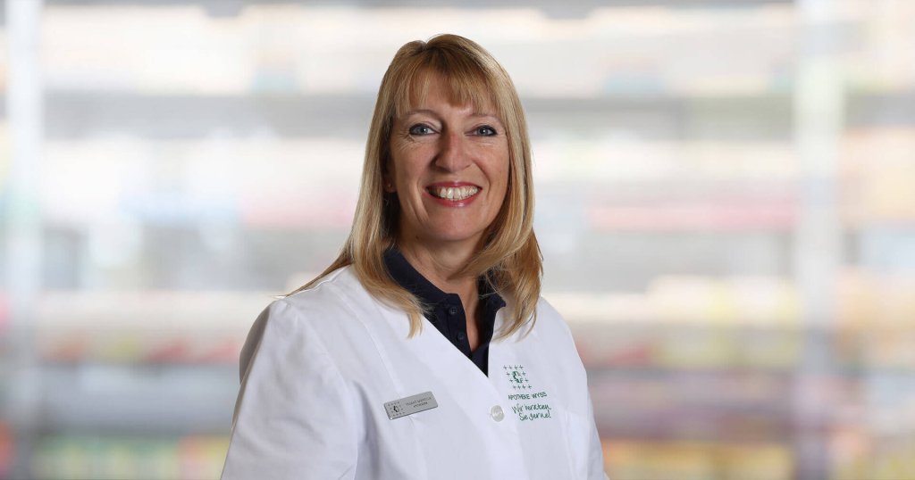 Gabriella Pagano, eine lächelnde Frau mit blondem Haar, trägt einen weißen Laborkittel und ein Namensschild, während sie vor einem unscharfen Innenhintergrund steht.