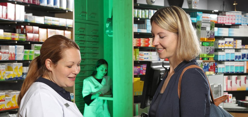 Eine Apothekerin im weißen Kittel berät eine lächelnde Kundin an der Apothekentheke über die Gesundheitsprodukte von Reise, während eine andere Mitarbeiterin zwischen den Regalen mit den verschiedenen Gesundheitsprodukten arbeitet.