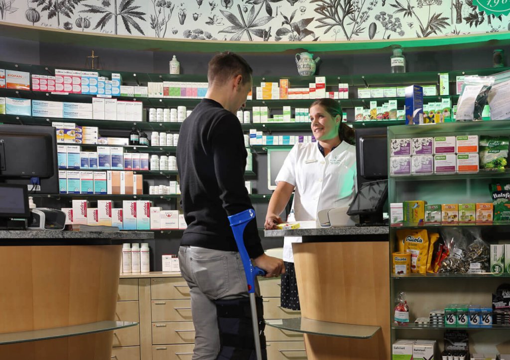 Ein Mann mit Krücken steht an einem Apothekentresen und spricht mit einem lächelnden Apotheker in einem weißen Kittel. Die Regale hinter dem Tresen sind mit Medikamenten, Gesundheitsprodukten und Sanitätsartikeln gefüllt.