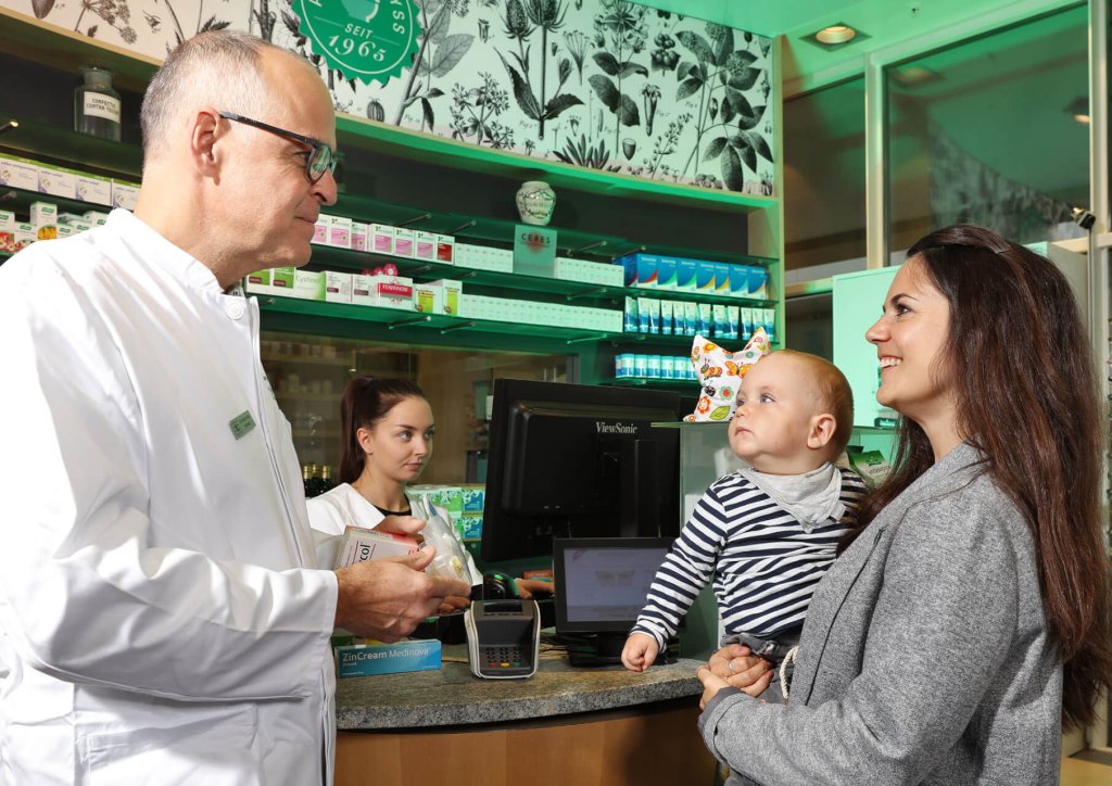 Ein Apotheker in einem wei&szlig;en Kittel spricht in einer Apotheke mit einer l&auml;chelnden Frau, die ein Baby h&auml;lt. Ein anderer Angestellter arbeitet am Computer hinter dem Tresen. Im Hintergrund sind Regale mit Medikamenten zu sehen.