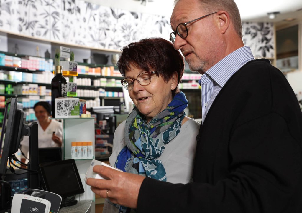 Eine &auml;ltere Frau und ein &auml;lterer Mann stehen zusammen in einer Apotheke und schauen auf eine Rezeptflasche. Im Hintergrund sind Regale mit Medikamenten zu sehen, und hinter dem Tresen arbeitet ein Apotheker.