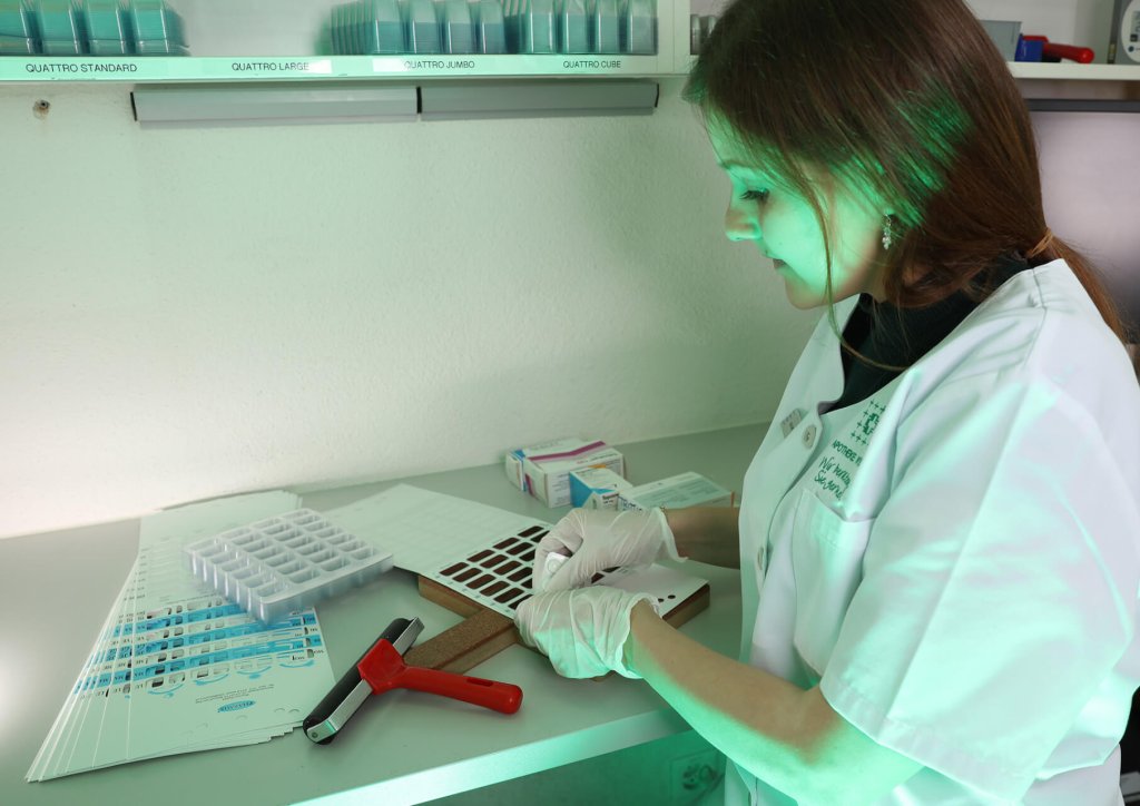 Eine Frau in Laborkittel und Handschuhen untersucht Proben an einem Laborarbeitsplatz, benutzt Werkzeuge und dokumentiert die Ergebnisse. Auf dem Schreibtisch sind Tabletts, ein Hammer, Formulare und ein Medikamenten-Dosiersystem unter gr&uuml;n get&ouml;ntem Licht zu sehen.