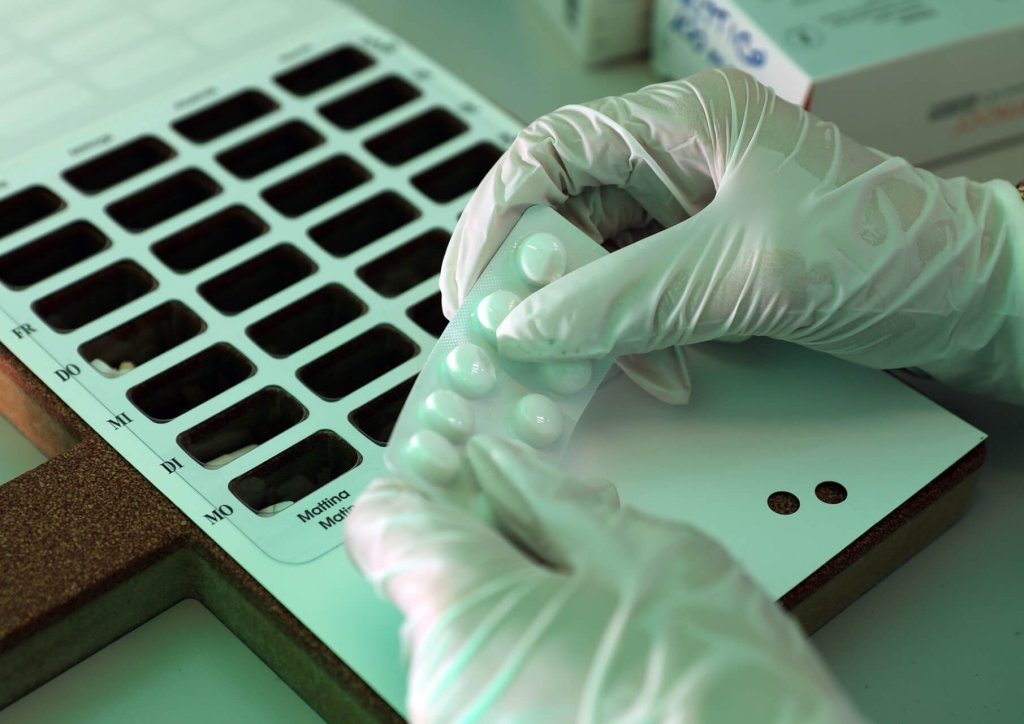 Eine Person mit wei&szlig;en Handschuhen h&auml;lt eine Blisterpackung mit Tabletten &uuml;ber ein Medikamenten-Dosiersystem mit beschrifteten F&auml;chern. Im Hintergrund sind Schachteln mit Medikamenten zu sehen.