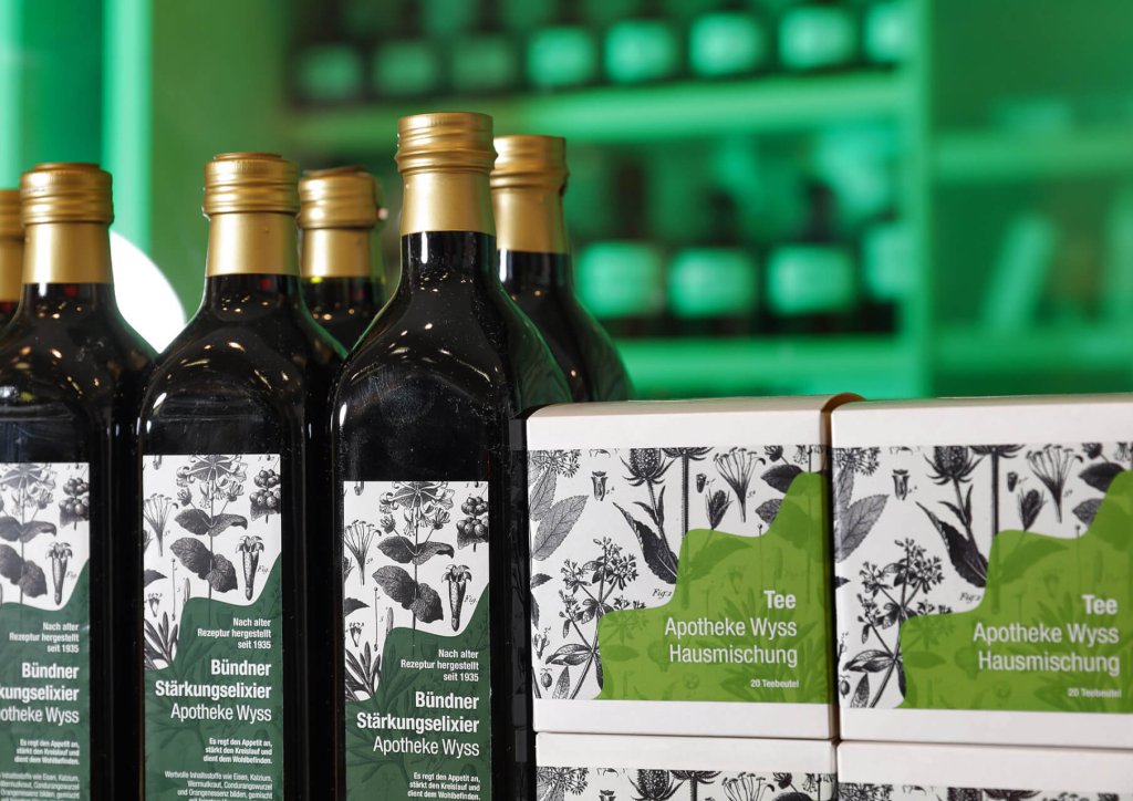 Mehrere dunkle Glasflaschen mit goldenen Verschl&uuml;ssen und gr&uuml;n-wei&szlig;en botanischen Etiketten stehen neben gr&uuml;nen Schachteln mit der Aufschrift "Tee Apotheke Wyss Hausmischung" der Wyss Manufaktur, alles vor einem verschwommenen gr&uuml;nen Hintergrund.