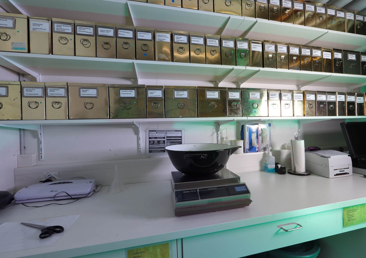 Ein Laborarbeitsplatz mit einer Digitalwaage, einer schwarz-weißen Schüssel, Scheren, Papieren und verschiedenen Geräten auf dem Tresen. Darüber befinden sich Regale mit vielen beschrifteten Metallbehältern mit runden Griffen.