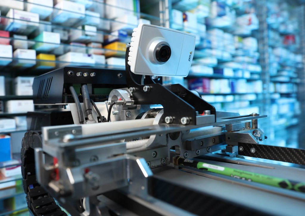 Nahaufnahme eines automatisierten Roboters mit Kamera in einer Apotheke am Standort Baden, umgeben von Regalen, die mit verschiedenen Medikamentenschachteln gef&uuml;llt sind und die fortschrittliche Technologie in der pharmazeutischen Automatisierung illustrieren.