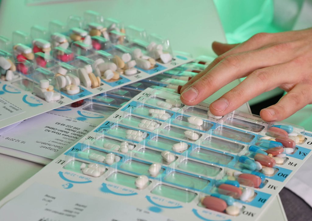 Eine Hand sortiert Blisterpackungen mit verschiedenfarbigen Pillen auf beschrifteten Medikamentenkarten, die nach einem Medikamenten-Dosiersystem für bestimmte Dosen oder Wochentage geordnet sind.