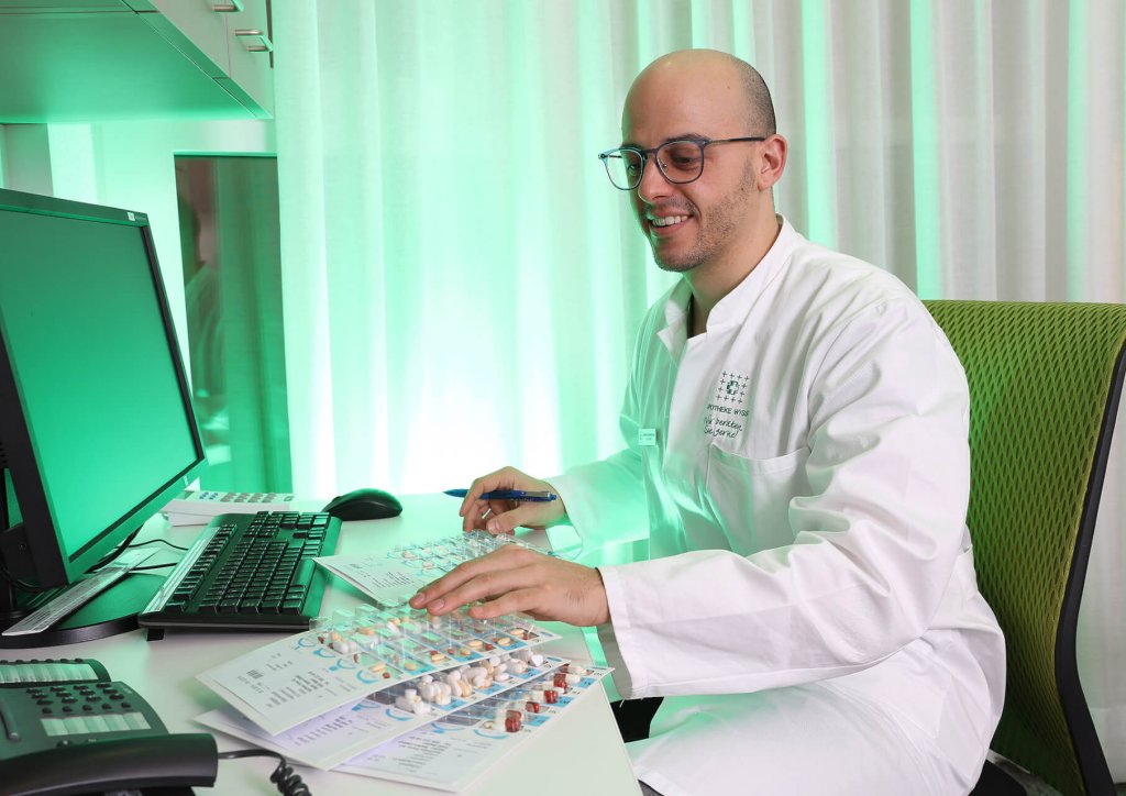 Eine Person in einem wei&szlig;en Laborkittel sitzt an einem Schreibtisch, l&auml;chelt und betrachtet Zahnmodelle und Krankenbl&auml;tter. Auf dem Schreibtisch in einem hell erleuchteten B&uuml;ro stehen ein Computer, Papierkram und eine Dosierhilfe f&uuml;r eine pr&auml;zise Medikamentendosierung.