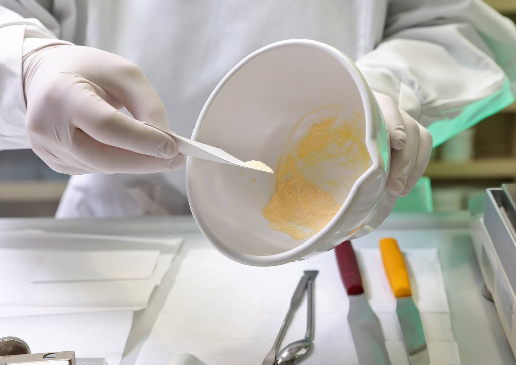 Eine Person mit wei&szlig;en Handschuhen und einem Laborkittel mischt mit einem Spatel ein gelbes Pulver in einer wei&szlig;en Sch&uuml;ssel. Auf der makellosen weissen Arbeitsfl&auml;che liegen verschiedene Werkzeuge und Utensilien der Wyss Manufaktur bereit.