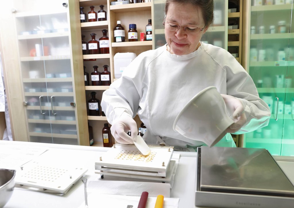 Eine Frau in Laborkittel und Handschuhen bereitet in einem Labor der Wyss Manufaktur mit M&ouml;rser und St&ouml;&szlig;el Kapseln vor. Hinter ihr stehen Flaschen mit Chemikalien und Ger&auml;te in Regalen.