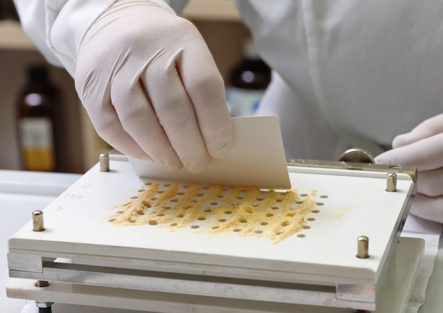 Eine Person mit weissen Handschuhen verteilt mit einer Karte gleichm&auml;ssig gelbes Pulver auf einer Kapself&uuml;llschale der Wyss Manufaktur in einer Laborumgebung.