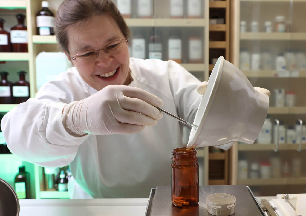 Eine l&auml;chelnde Frau in wei&szlig;em Laborkittel und Handschuhen sch&uuml;ttet im Labor der Wyss Manufaktur mit einem Spatel Pulver aus einer Schale in ein braunes Glasgef&auml;&szlig;, im Hintergrund stehen Regale mit Flaschen.