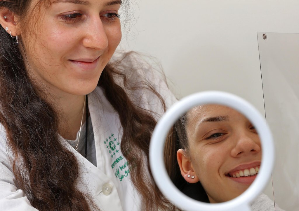 Eine l&auml;chelnde Frau in einem wei&szlig;en Kittel betrachtet in einem runden Spiegel das Spiegelbild einer anderen Frau, die ebenfalls l&auml;chelt, in einer hellen, klinischen Umgebung - perfekt f&uuml;r Momente wie Ohrloch stechen.