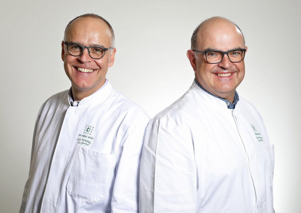 Zwei Männer mittleren Alters mit Brille und kurzen Haaren, beide in weißen Laborkitteln, stehen nebeneinander und lächeln in die Kamera vor einem schlichten, hellen Hintergrund.