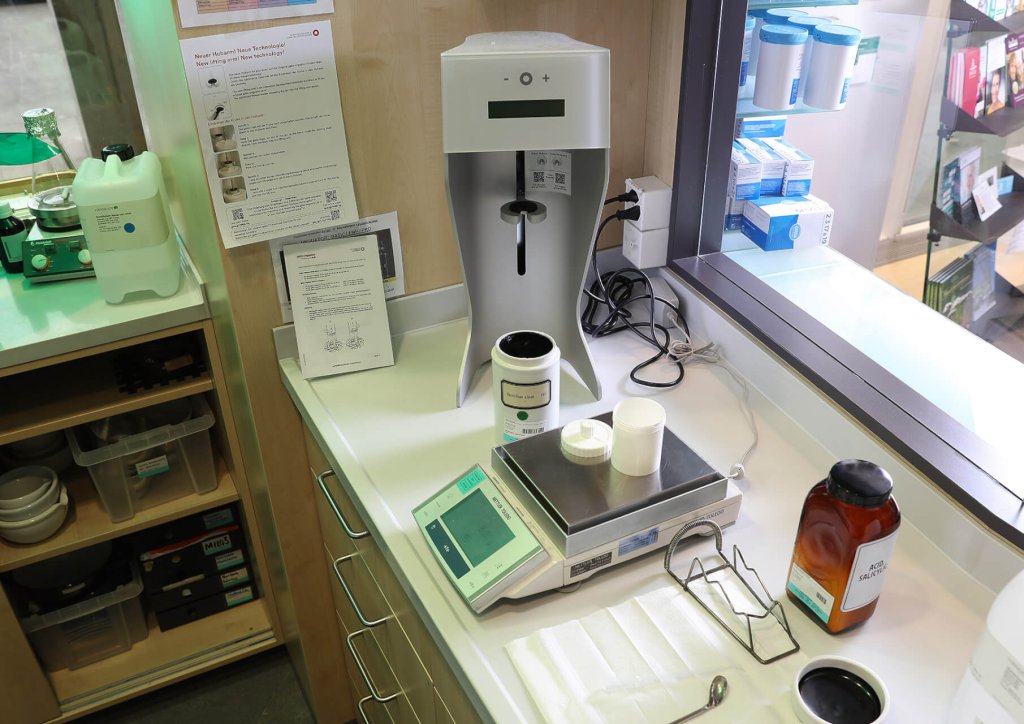 Ein Apotheken-Compoundier-Arbeitsplatz der Wyss Manufaktur mit einer Digitalwaage, einem Pulverbeh&auml;lter, einer Bernsteinflasche, Papierb&ouml;gen und anderen Ger&auml;ten auf einem weissen Tresen in der N&auml;he von Regalen mit pharmazeutischem Bedarf.