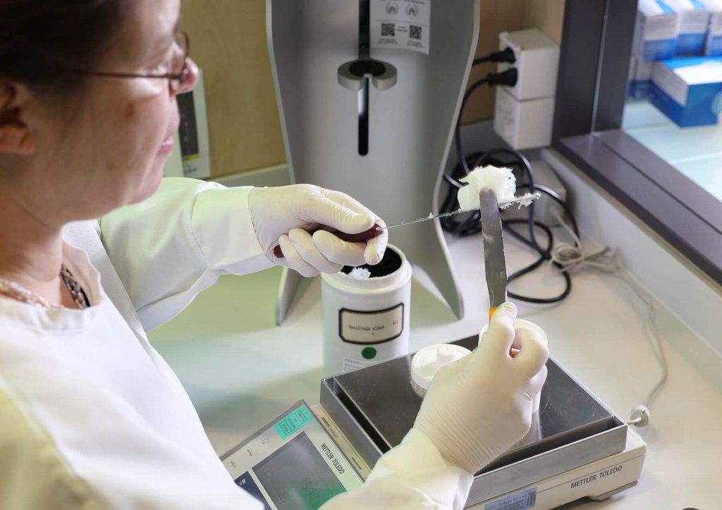 Ein Labortechniker der Wyss Manufaktur, der Handschuhe tr&auml;gt, benutzt einen Spatel und eine Schaufel, um eine weisse, flauschige Substanz &uuml;ber einer digitalen Waage in einer Laborumgebung zu handhaben.