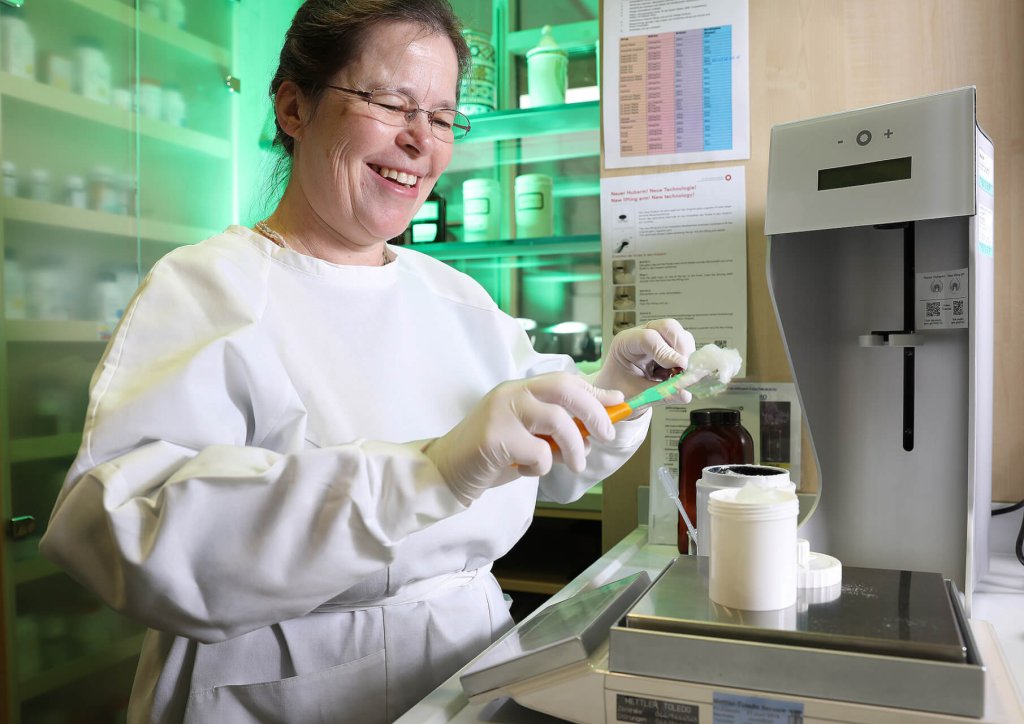 Eine l&auml;chelnde Frau in einem wei&szlig;en Laborkittel und mit Handschuhen bereitet in einem Labor der Wyss Manufaktur eine Probe vor. Sie benutzt einen Spatel, um eine Substanz in einen Beh&auml;lter auf einer digitalen Waage zu f&uuml;llen, w&auml;hrend im Hintergrund Laborger&auml;te und Gl&auml;ser zu sehen sind.