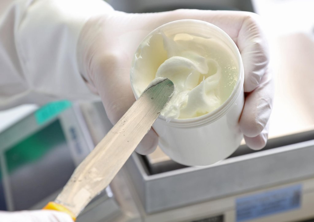 Eine Person mit wei&szlig;en Handschuhen h&auml;lt ein ge&ouml;ffnetes wei&szlig;es Cremet&ouml;pfchen, aus dem sie mit einem Metallspatel etwas herausl&ouml;ffelt. Die Laborausr&uuml;stung im Hintergrund deutet auf eine klinische oder pharmazeutische Umgebung der Wyss Manufaktur hin.