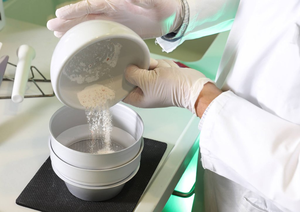 Eine Person, die einen Laborkittel und Handschuhe trägt, schüttet ein weißes Pulver aus einer Schale in einen Stapel von Sieben auf einem Labortisch der Wyss Manufaktur, wahrscheinlich zur Vorbereitung eines wissenschaftlichen Tests oder einer Qualitätskontrolle.