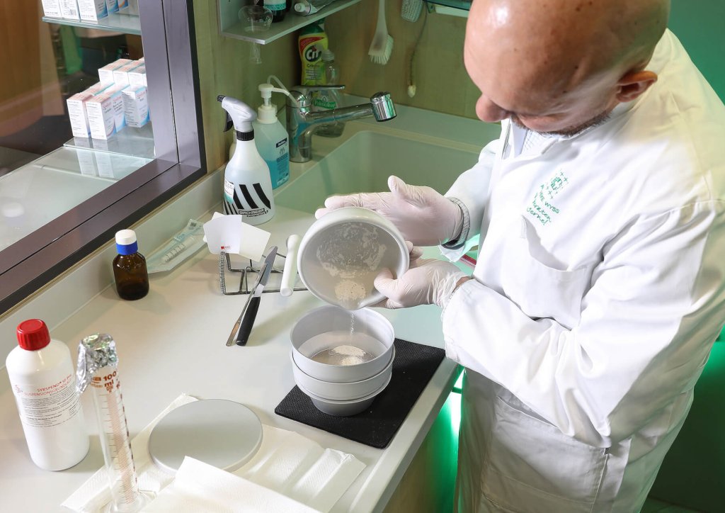 Eine Person in wei&szlig;em Laborkittel und Handschuhen sch&uuml;ttet in einem Labor der Wyss Manufaktur Pulver in einen M&ouml;rser. Auf dem Tresen stehen Laborger&auml;te, Flaschen und Reinigungsmittel.