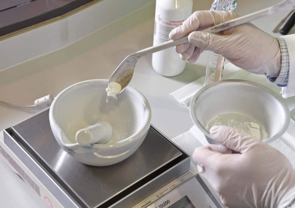 Eine Person mit Handschuhen f&uuml;llt mit einem L&ouml;ffel wei&szlig;es Pulver aus einer Schale in einen M&ouml;rser, der auf einer elektronischen Waage im Labor der Wyss Manufaktur steht.