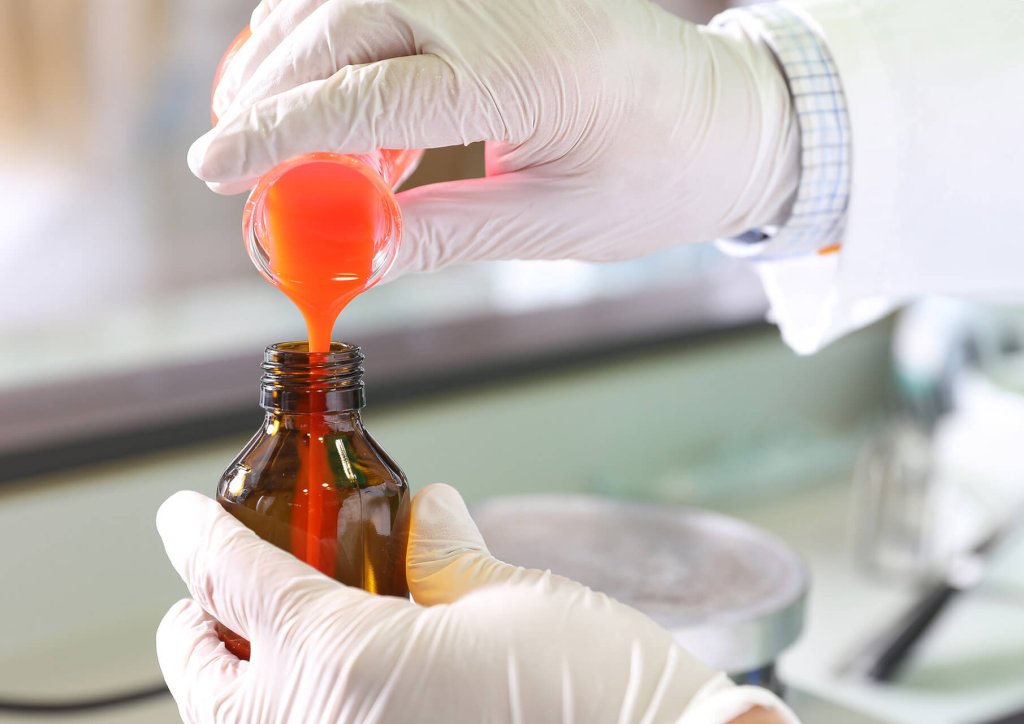 Eine Person mit wei&szlig;en Handschuhen gie&szlig;t eine orangefarbene Fl&uuml;ssigkeit aus einem Becherglas in eine braune Glasflasche, wahrscheinlich in einer Laborumgebung der Wyss Manufaktur.