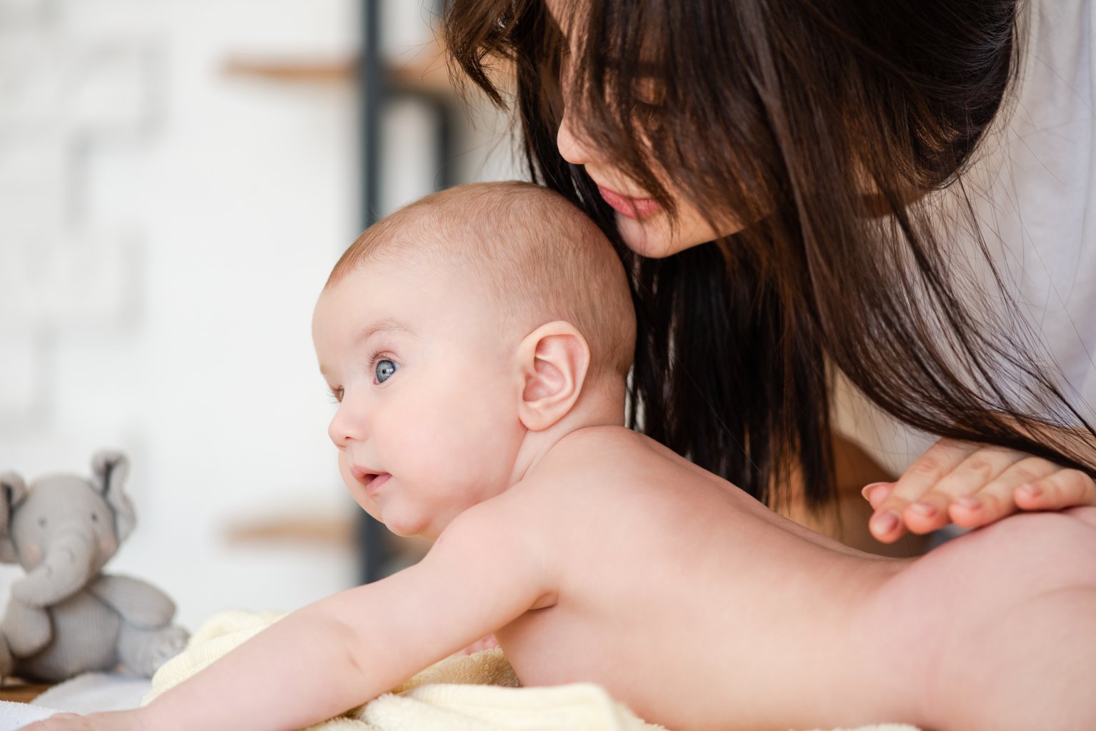 Eine Frau mit langen braunen Haaren beugt sich sanft über ein nacktes Baby, das auf einem weichen gelben Handtuch liegt, und stützt es, während es mit großen blauen Augen nach vorne schaut. Die Szene ist hell und zart.