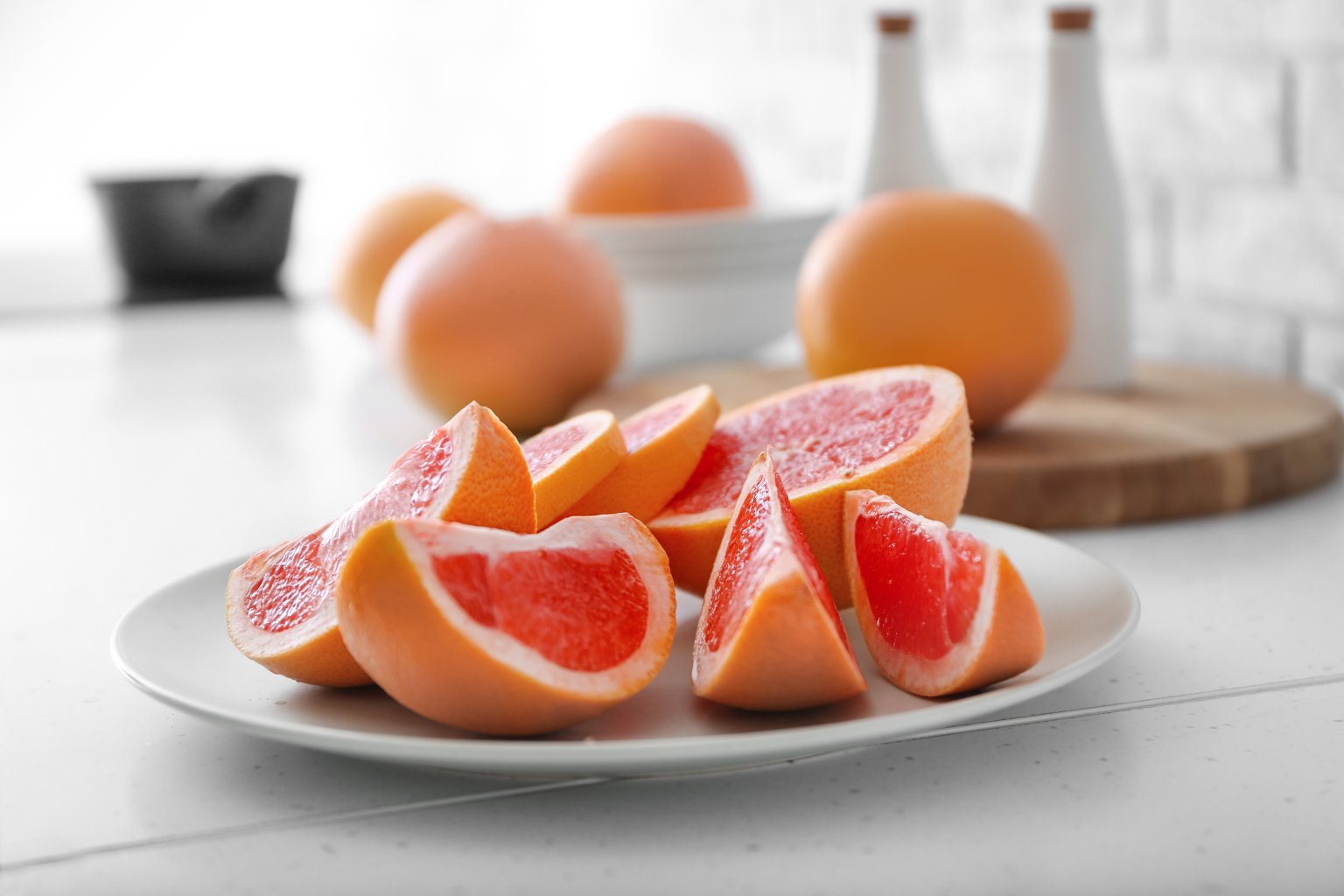 Aufgeschnittene Grapefruitscheiben auf einem wei&szlig;en Teller, mit ganzen Grapefruits und einem h&ouml;lzernen Schneidebrett im Hintergrund auf einer hellen K&uuml;chenzeile.